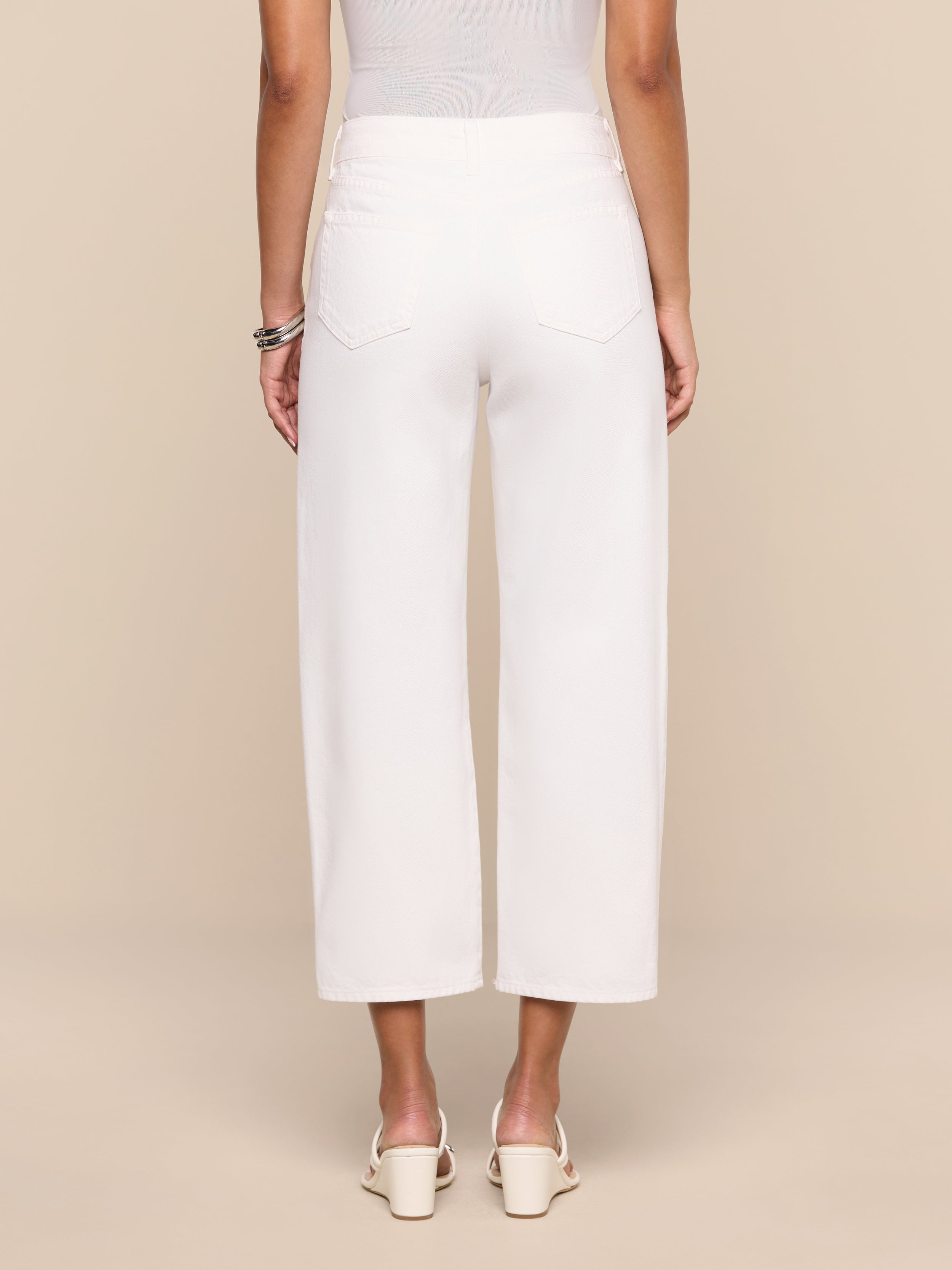 A person stands facing away from the camera, wearing the Liora Cropped Jean in white—a high rise, wide-leg style—paired with a sleeveless top, white heeled shoes, and bracelets, against a plain white background.