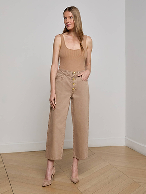 A woman with long straight hair stands indoors on a wooden floor, wearing L'AGENCE's Liora Cropped Jean and a sleeveless tan top, paired with tan heels. She poses against a plain wall.