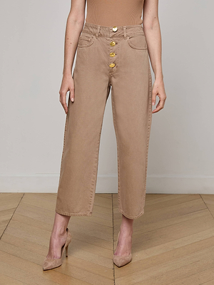 A woman stands on a wooden floor wearing L'AGENCE's Liora Cropped Jean, a high-rise, wide-leg beige jean with large gold buttons, paired with a beige sleeveless top and matching beige heels. Only her body from the shoulders down is visible.