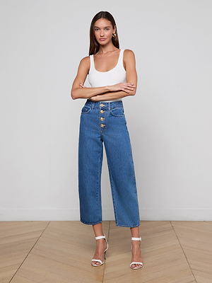 A woman stands with arms crossed, wearing a white tank top and the Liora Cropped Jean—high rise blue wide leg cropped jeans with gold buttons—paired with white high-heeled sandals, posing indoors against a light wall and wooden floor.
