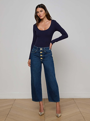 A woman poses indoors against a plain wall, wearing the L’AGENCE Liora Cropped Jean—a high-rise, wide-leg blue denim with large gold buttons—paired with a fitted navy long-sleeve top and metallic pointed-toe heels.
