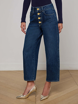 Wearing L'AGENCE Liora Cropped Jeans—high rise, wide leg blue denim with four large gold buttons—a person pairs them with a fitted dark long-sleeve top and shiny gold pointed-toe heels, standing on a wooden floor against a plain wall.