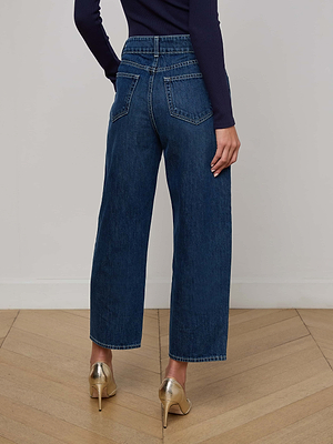 Someone wearing L'AGENCE Liora Cropped Jeans, a long-sleeve navy top, and gold high-heeled shoes stands on a wooden floor facing away from the camera.