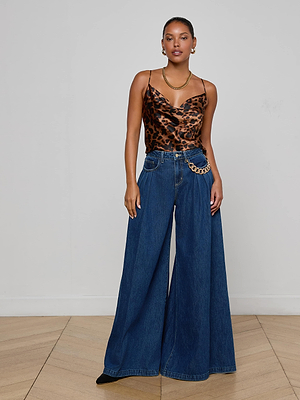 A woman poses indoors on a wooden floor, wearing L'AGENCE's Chayse Wide-Leg Jean in non-stretch denim with a statement chain belt, a leopard print camisole, gold hoop earrings, and a necklace against a plain light background.