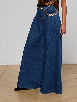 A person wearing the Chayse Wide-Leg Jean by L’AGENCE with pleats and a statement waist chain, paired with a leopard print sleeveless top, is standing on a wooden floor.