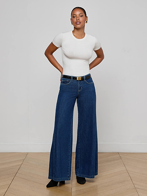 A woman poses confidently against a white wall, wearing a fitted white short-sleeve top and L’AGENCE Savina Low-Rise Wide-Leg Jeans in indigo wash, paired with a black belt and shoes. Her hands are behind her back as she looks at the camera.