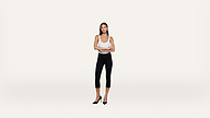 A woman stands confidently with arms crossed, wearing a white tank top, black fitted pants, and black heels, against a plain light background.