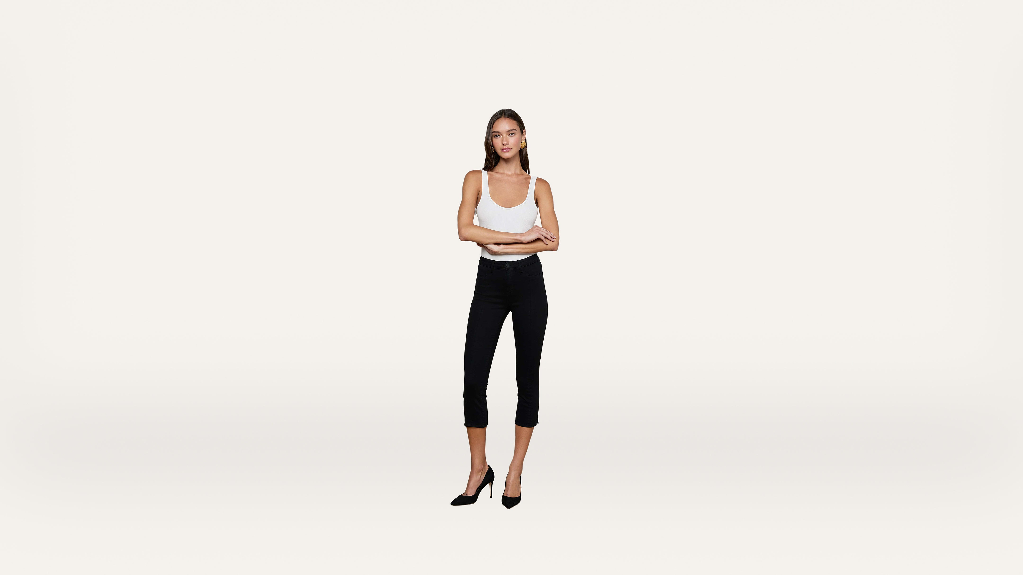 A woman stands confidently with arms crossed, wearing a white tank top, black fitted pants, and black heels, against a plain light background.