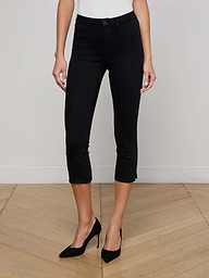 A person wearing L'AGENCE Fiora Pedal Pusher Jeans in black and black high heels stands on a light wood herringbone floor against a plain light-colored wall; their upper body is not visible.