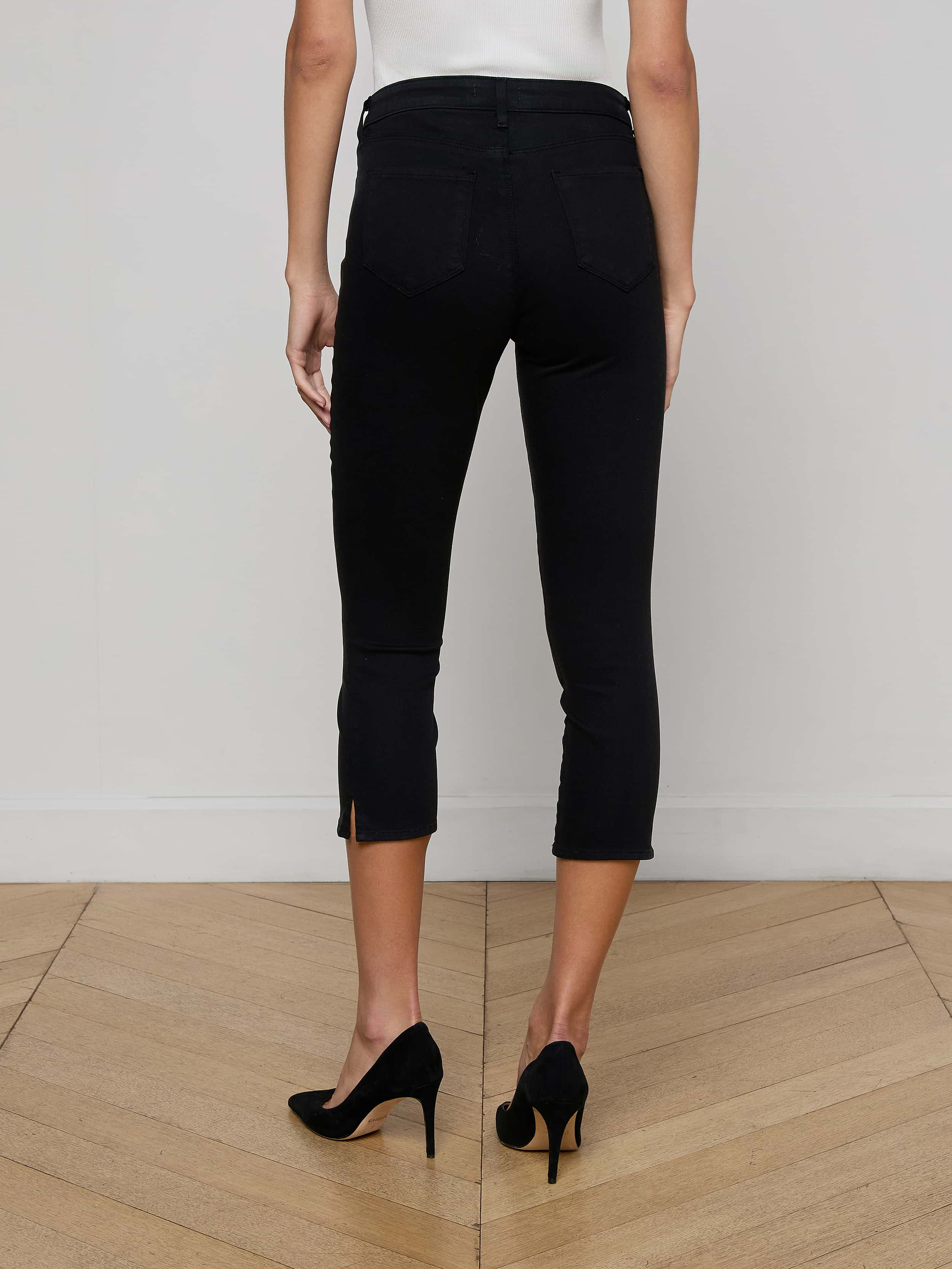 A woman stands on a wooden floor facing away, wearing L'AGENCE Fiora Pedal Pusher Jeans with back pockets, a white sleeveless top, and black high-heeled shoes.