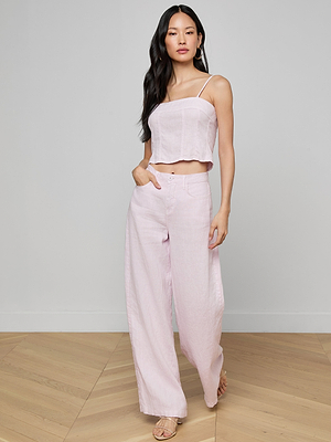A woman with long dark hair stands indoors, looking confidently at the camera with one hand in her pocket, wearing a light pink sleeveless cropped top and the Amara Linen Wide-Leg Pant.