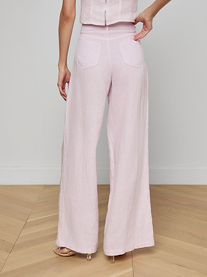 A person stands on a wood floor indoors, wearing the Amara Linen Wide-Leg Pant in light pink with a matching top. The image, taken from behind, showcases the pant’s back pockets and loose high-rise fit.