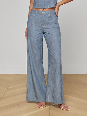 A woman wearing the Amara Wide-Leg Pant in blue and white pinstripes with a matching crop top and beige heeled sandals stands on a light wood floor against a plain white wall.