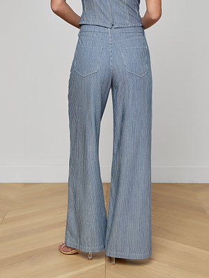 A person stands with their back to the camera, wearing the Amara Wide-Leg Pant in blue and white indigo stripes, paired with heeled sandals on a light wood floor. The upper body is partially visible.