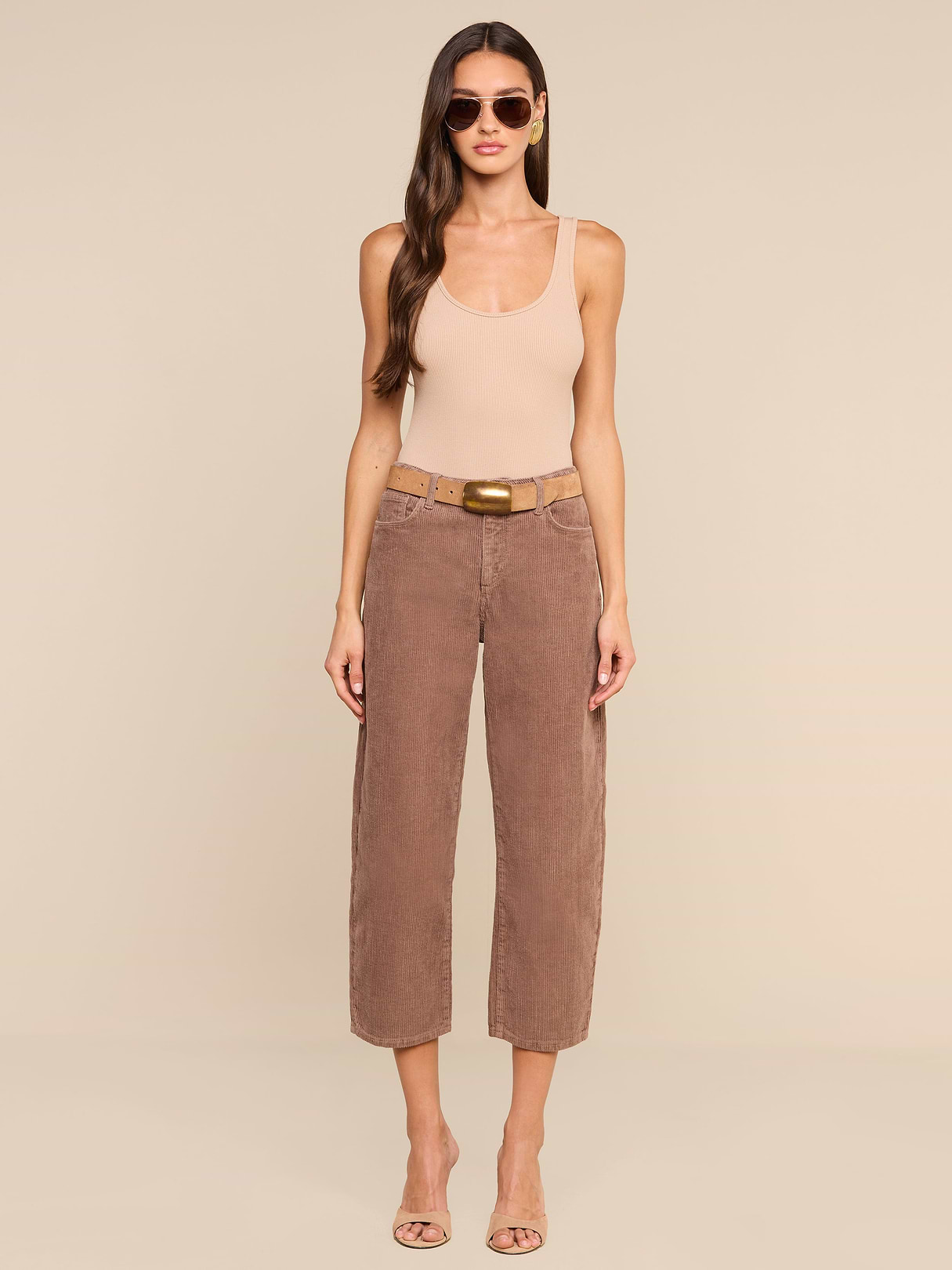 A woman stands against a beige background, wearing sunglasses and a sleeveless beige top paired with the Mallis Corduroy Bow-Leg Pant—a high-rise, brown cropped style featuring a tan belt—perfect for elevating any casual wardrobe. She completes the look with open-toed heels. Her long, wavy hair frames her relaxed silhouette, with arms at her sides.