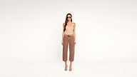 A woman wearing sunglasses, a beige tank top, brown wide-leg pants, and sandals stands facing forward against a plain white background.