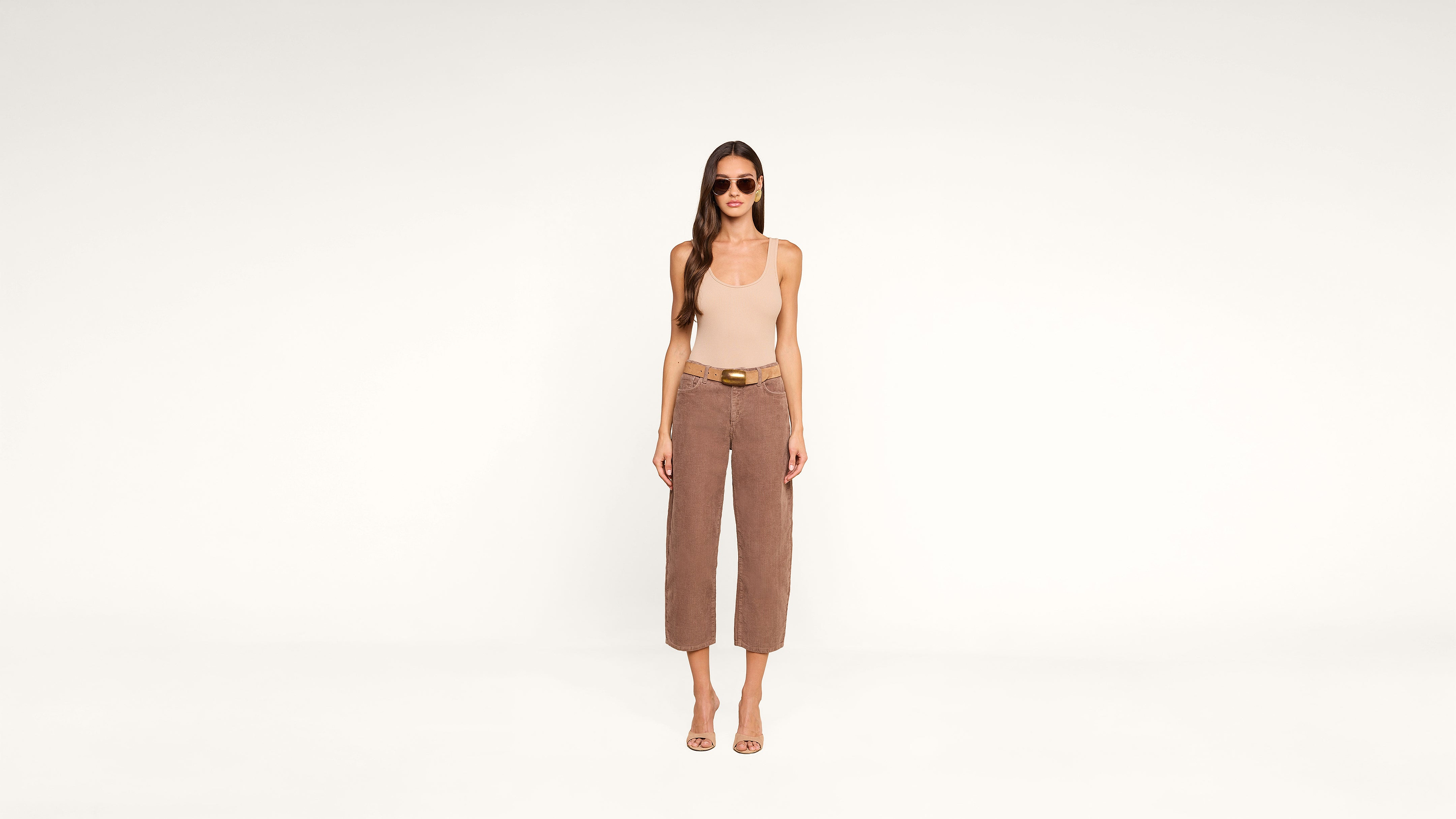 A woman wearing sunglasses, a beige tank top, brown wide-leg pants, and sandals stands facing forward against a plain white background.