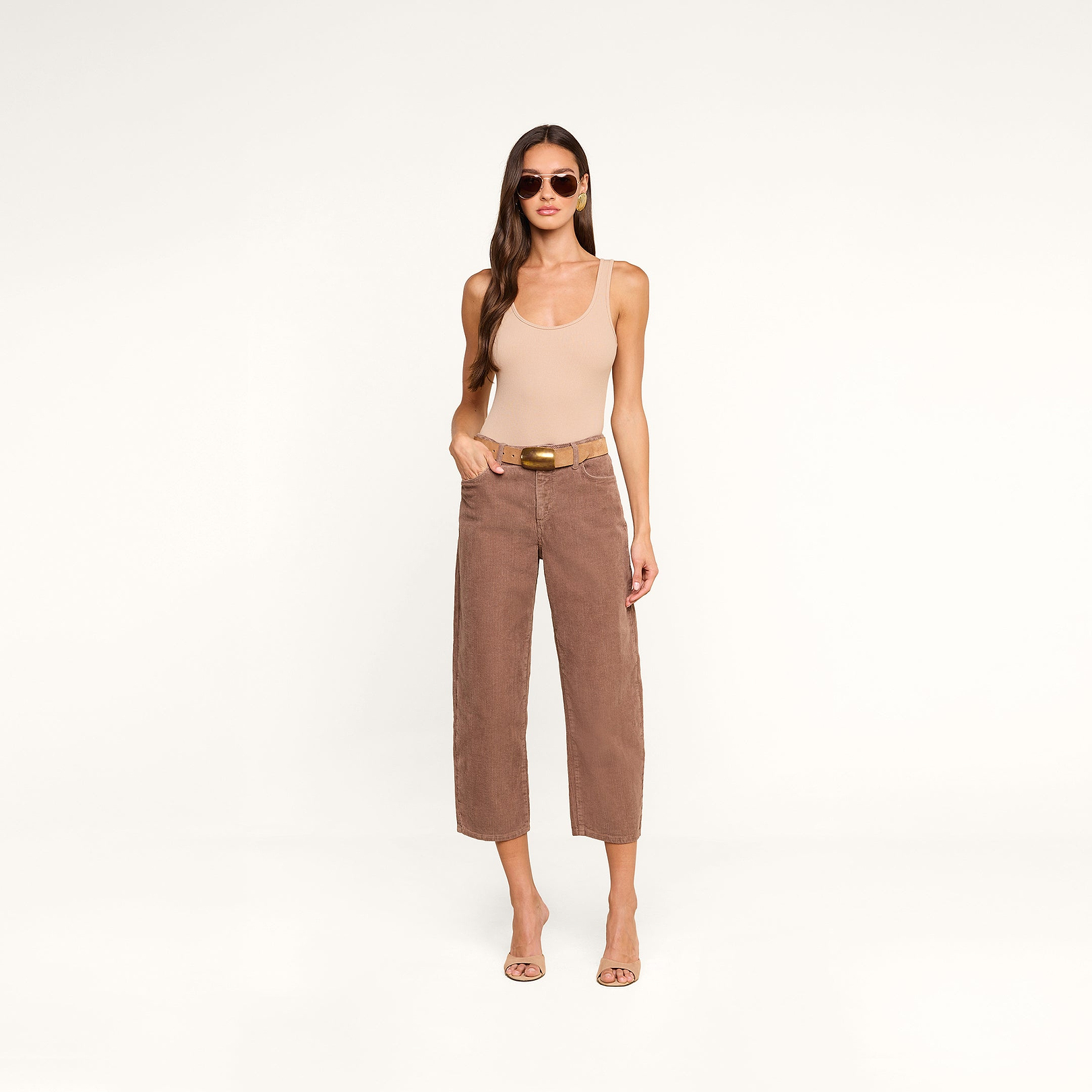 A woman wearing sunglasses, a nude tank top, and the Mallis Corduroy Bow-Leg Pant with a belt stands on a beige background, completing her casual look with heeled sandals.