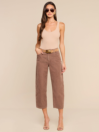 A woman wearing sunglasses, a nude tank top, and the Mallis Corduroy Bow-Leg Pant with a belt stands on a beige background, completing her casual look with heeled sandals.