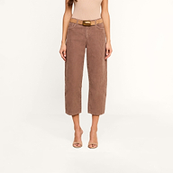 A woman wearing the Mallis Corduroy Bow-Leg Pant in brown, styled with a tan sleeveless top, an oversized belt featuring a gold buckle, and open-toed heels stands against a plain white background. Her head is not visible.