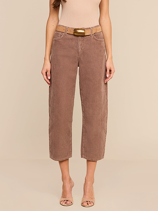 A woman stands against a plain beige background wearing the Mallis Corduroy Bow-Leg Pant in a rich brown shade, styled with a tan top, wide tan belt with a gold buckle, and beige open-toe heels—an effortlessly chic look perfect for any casual wardrobe.