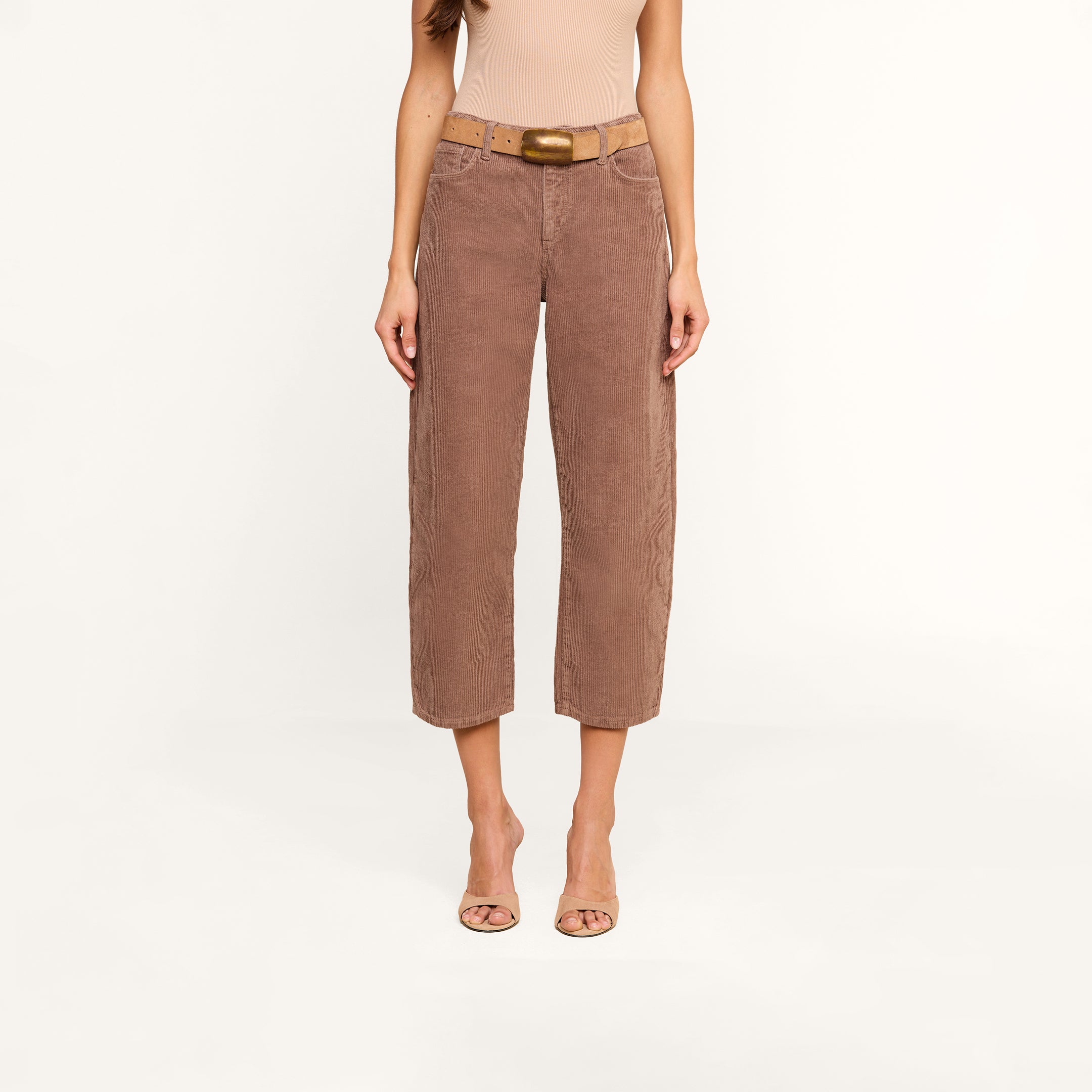 A woman wearing the Mallis Corduroy Bow-Leg Pant in brown, styled with a tan sleeveless top, an oversized belt featuring a gold buckle, and open-toed heels stands against a plain white background. Her head is not visible.