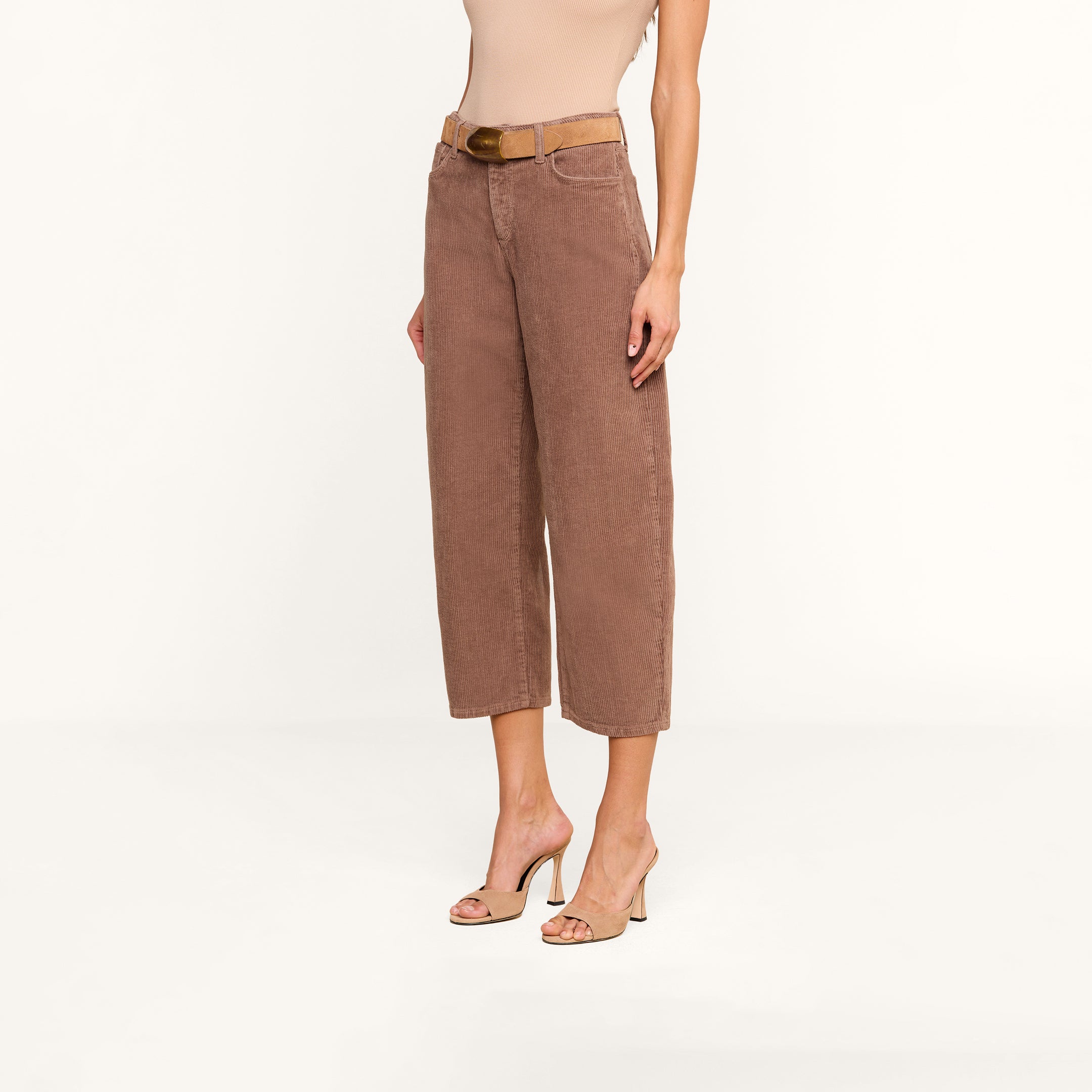 A person wearing the Mallis Corduroy Bow-Leg Pant in brown stands against a plain white background. They have paired the high-rise, wide-leg corduroy pants with a beige belt, a sleeveless beige top, and beige heeled sandals. Their face is not visible.