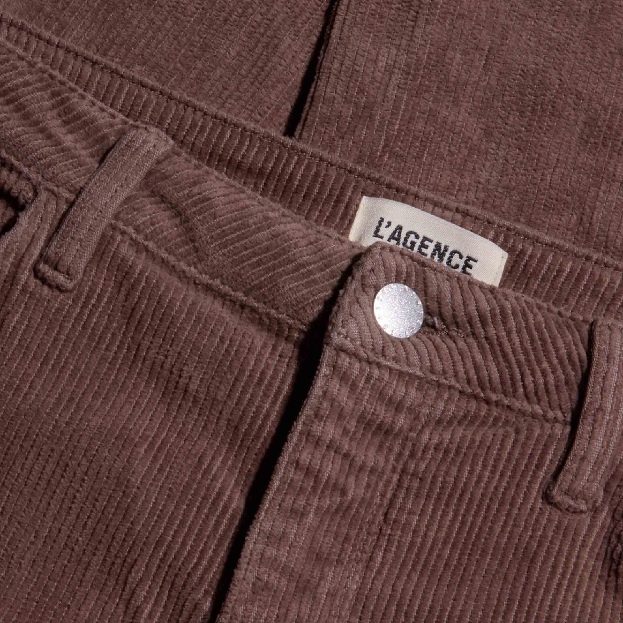 Close-up of the Mallis Corduroy Bow-Leg Pant in brown, featuring a high rise, visible button, front pocket, and a label reading LAGENCE above the waistband.