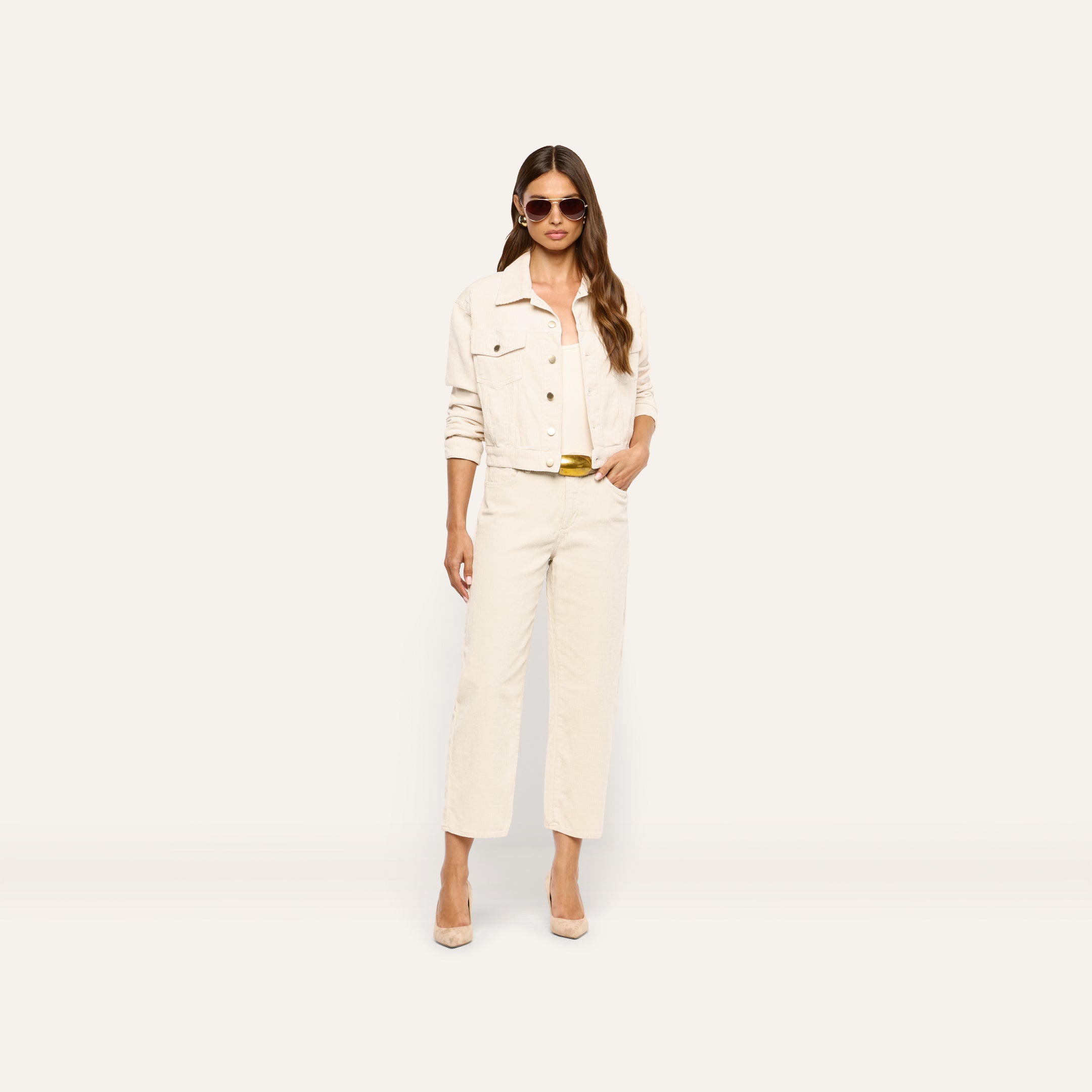 A person wears a cropped cream-colored jacket, matching tank top, and Mallis Corduroy Bow-Leg Pant with a gold-tone belt, paired with beige pumps and sunglasses.
