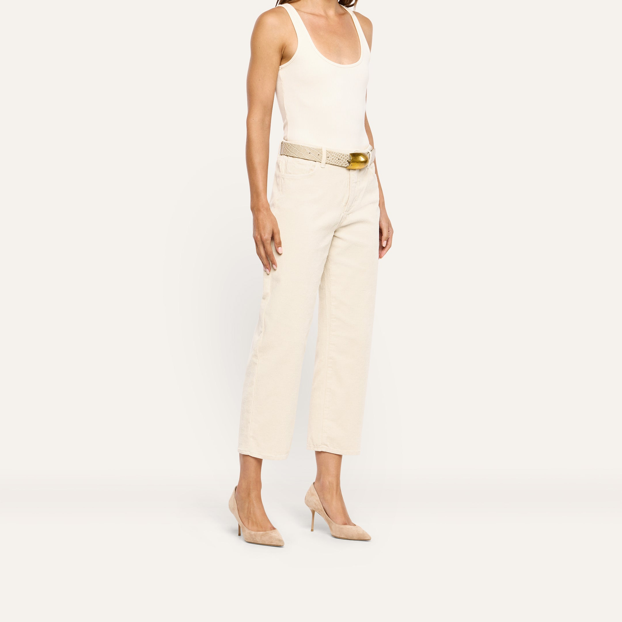 A person wears a white tank top, a cream-colored textured belt with a gold buckle, wide-leg high-rise Mallis Corduroy Bow-Leg Pant, and pointed-toe heels.
