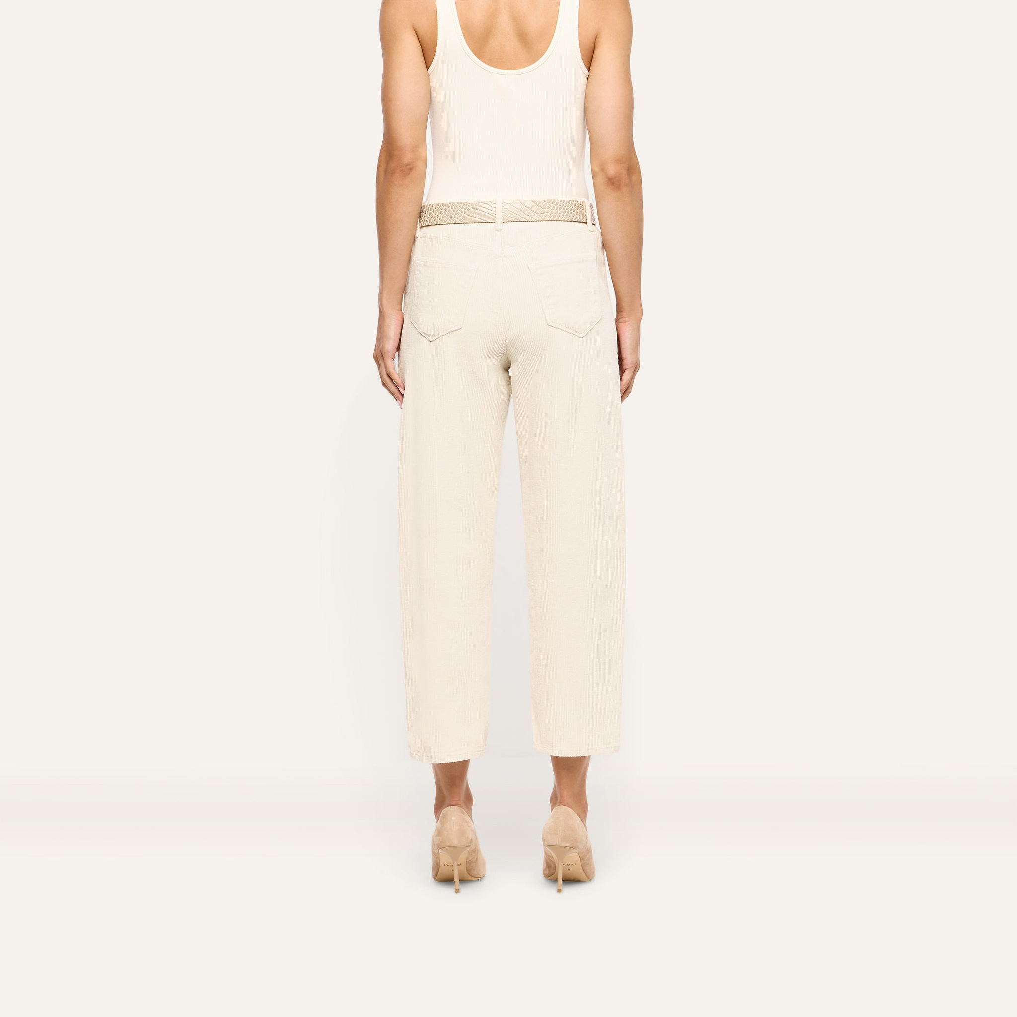 Back view of a person wearing Mallis Corduroy Bow-Leg pants in beige, featuring a high-rise, relaxed wide-leg silhouette and a woven belt, styled with a tank top and heeled pumps.