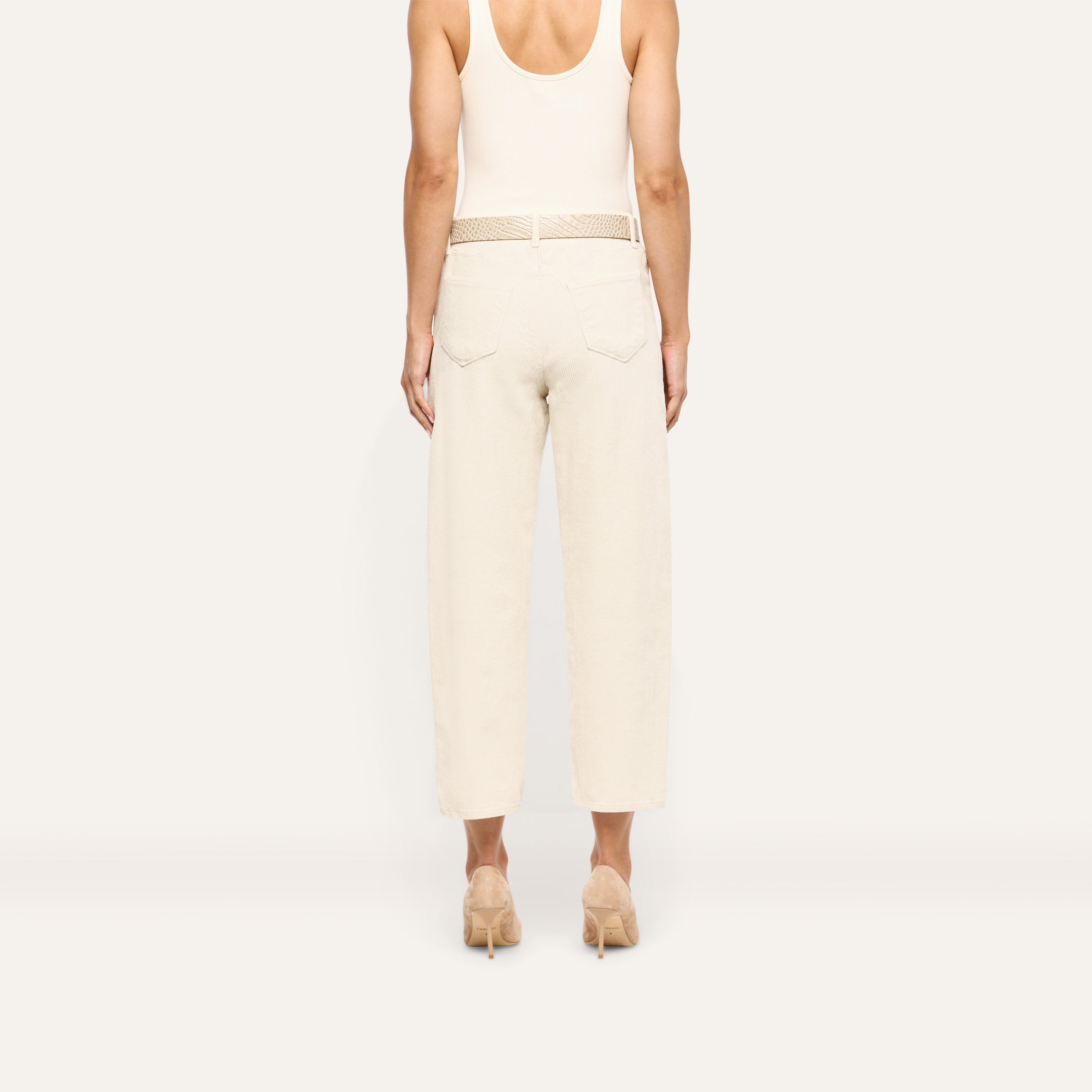 Back view of a person wearing Mallis Corduroy Bow-Leg pants in beige, featuring a high-rise, relaxed wide-leg silhouette and a woven belt, styled with a tank top and heeled pumps.