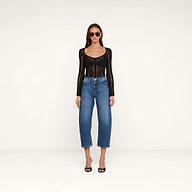 A woman wearing sunglasses, a black long-sleeve top, and black heels stands facing forward against a plain white background. She is dressed in the Mallis Bow-Leg Jean.