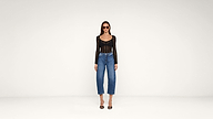 A woman wearing sunglasses, a black long-sleeve top, blue jeans, and black heels stands against a plain white background.