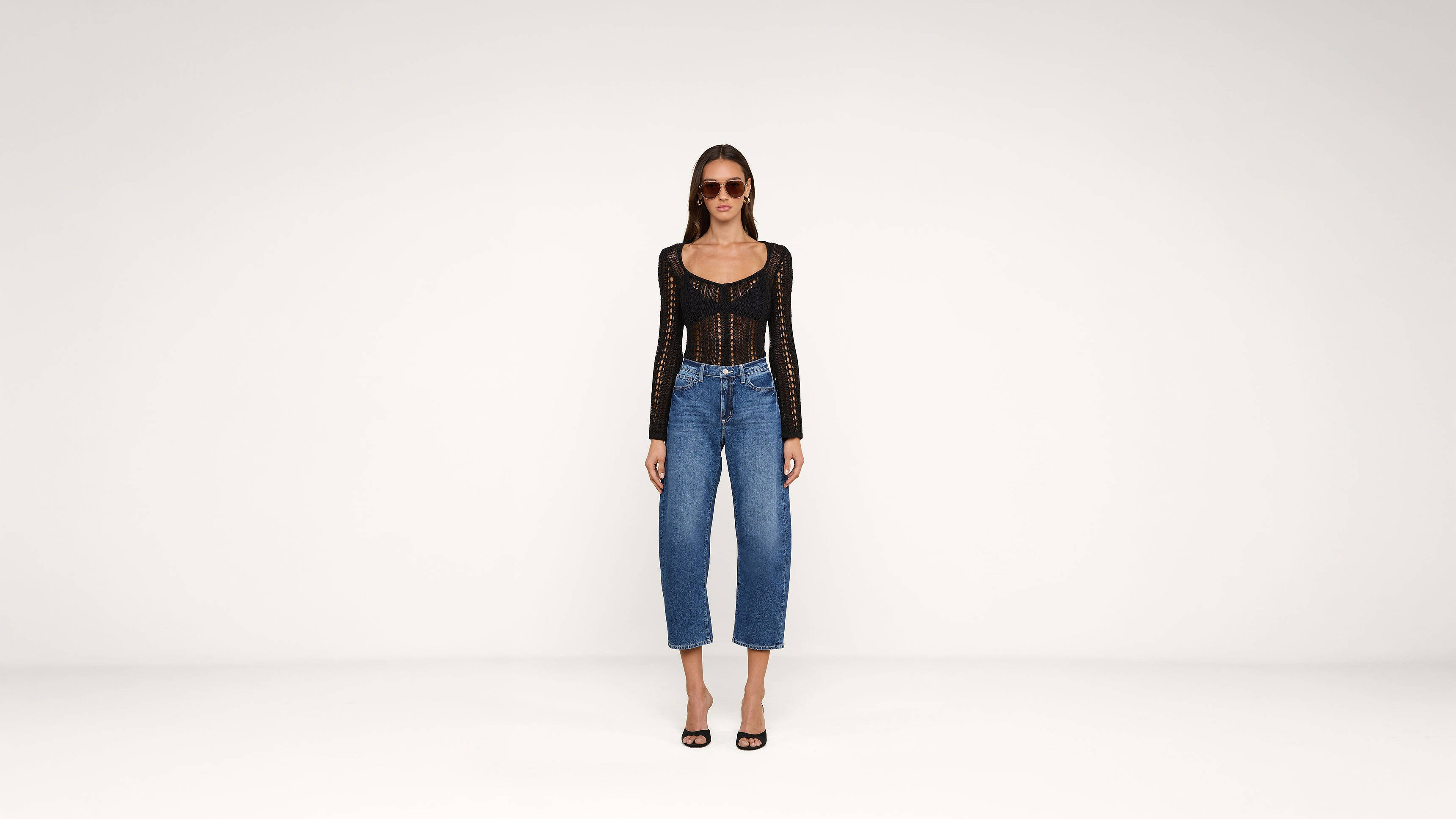 A woman wearing sunglasses, a black long-sleeve top, blue jeans, and black heels stands against a plain white background.