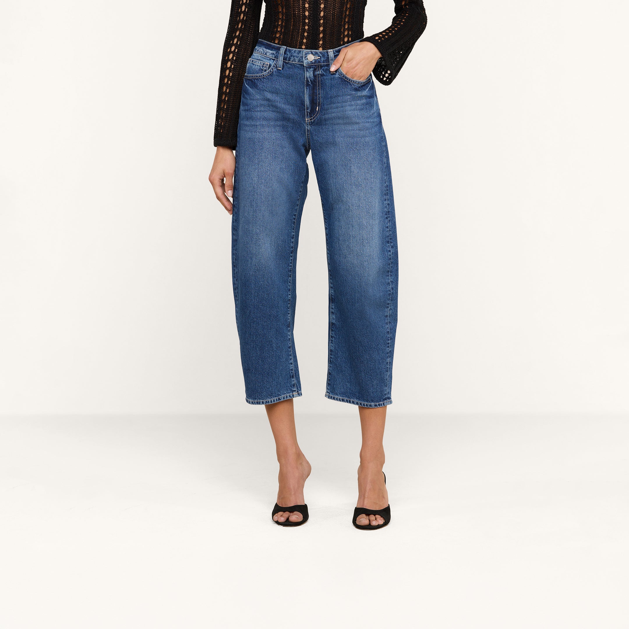 A person wearing the Mallis Bow-Leg Jean in denim blue, a black long-sleeve textured top, and black open-toe heels stands against a plain white background.