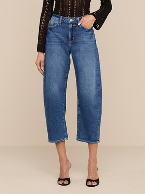 A person wears the Mallis Bow-Leg Jean, a high-waisted, wide-leg all-cotton denim, paired with a black long-sleeve top and black open-toe heels, standing against a beige background.
