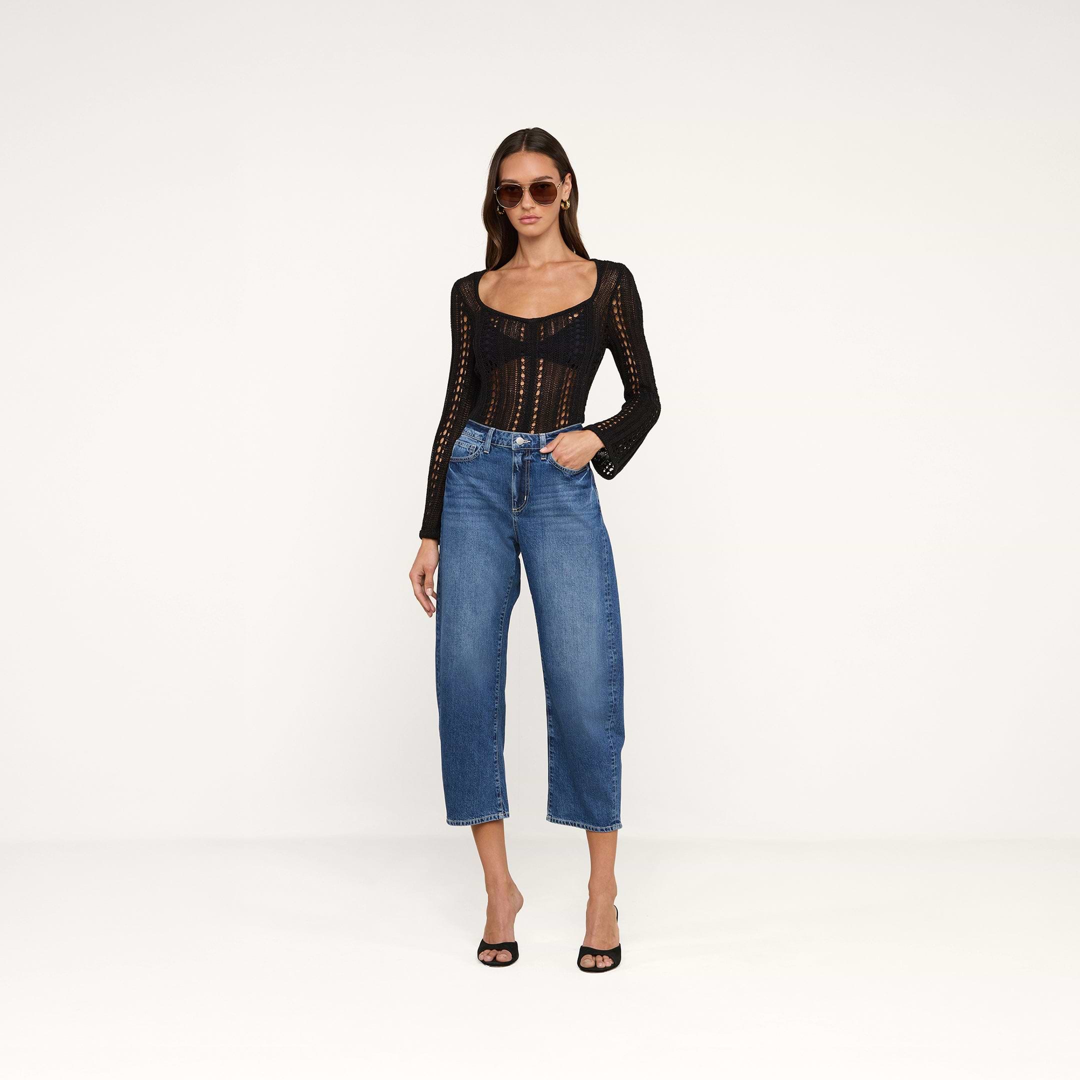 A woman wearing sunglasses, a black long-sleeve crochet top, the Mallis Bow-Leg Jean in blue high rise denim, and black heeled sandals stands against a plain white background.