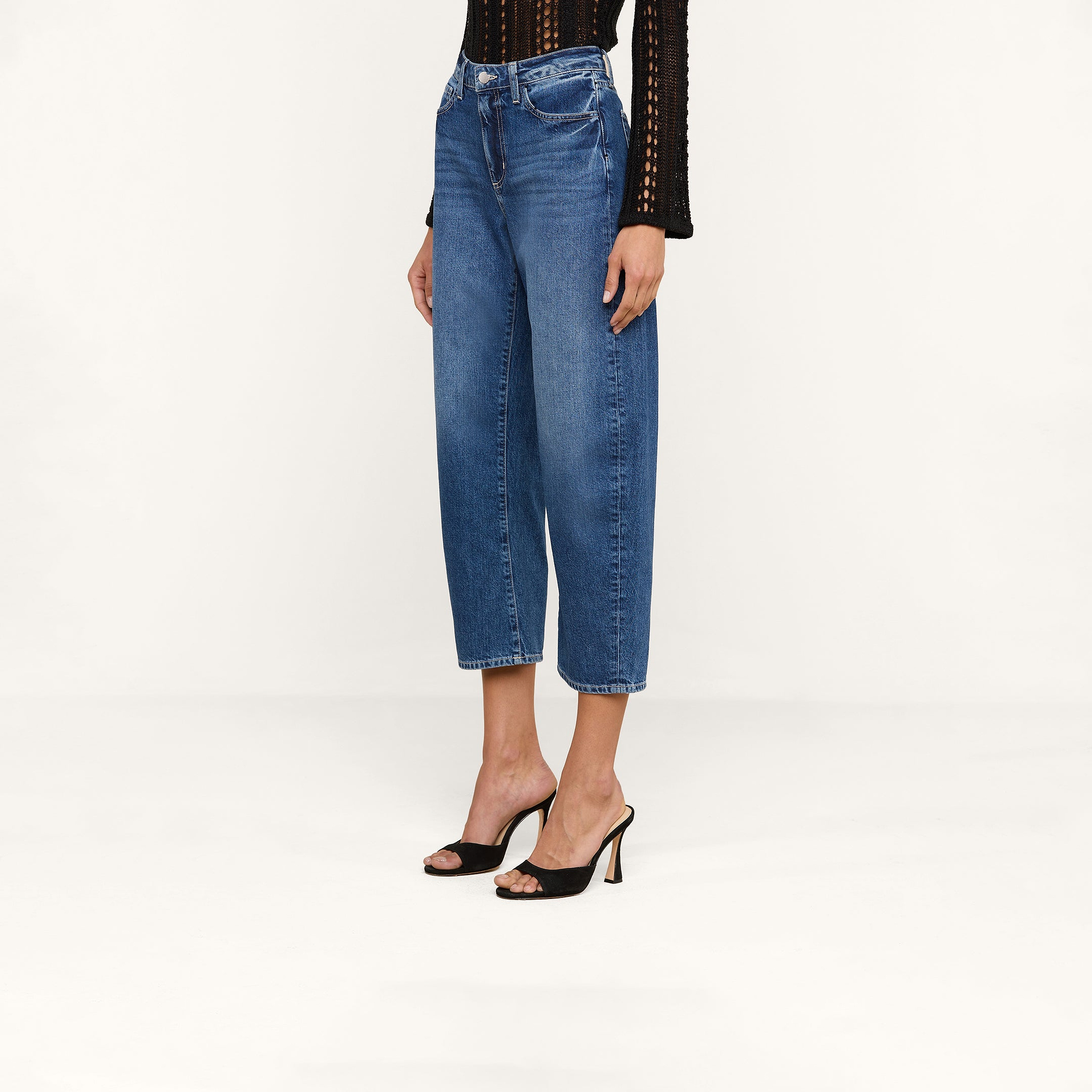Wearing the Mallis Bow-Leg Jean in a high-rise, cropped blue denim style, paired with a black long-sleeve top and black heeled sandals, a person stands against a plain white background.