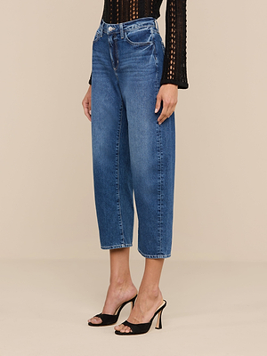 A person wearing Mallis Bow-Leg Jean high-rise, wide-leg blue jeans with a black textured long-sleeve top and black open-toe heeled sandals stands against a plain beige background.