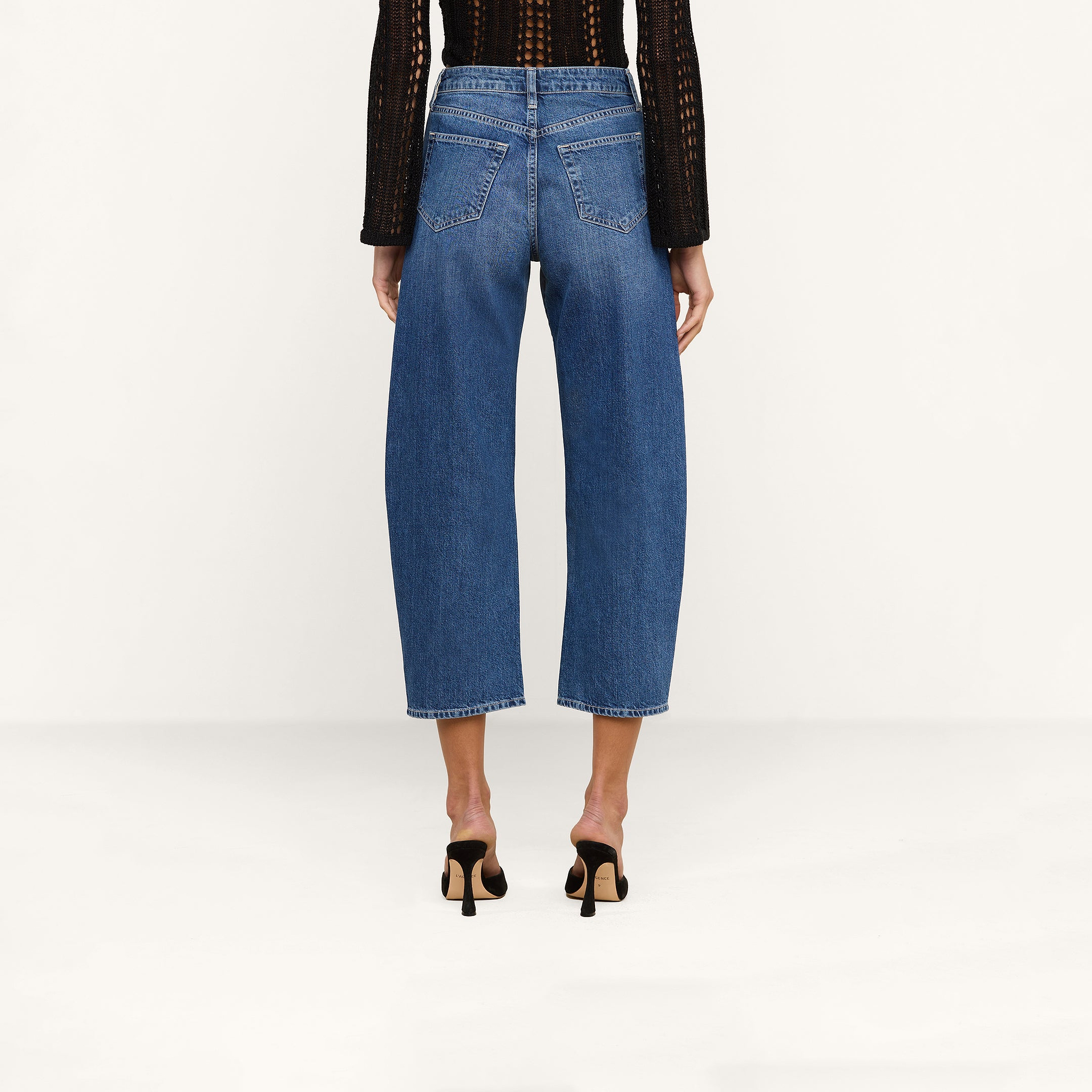 A person stands facing away against a plain white background, wearing the Mallis Bow-Leg Jean—a pair of blue high rise denim cropped jeans—styled with a black long-sleeved top and black heeled sandals.