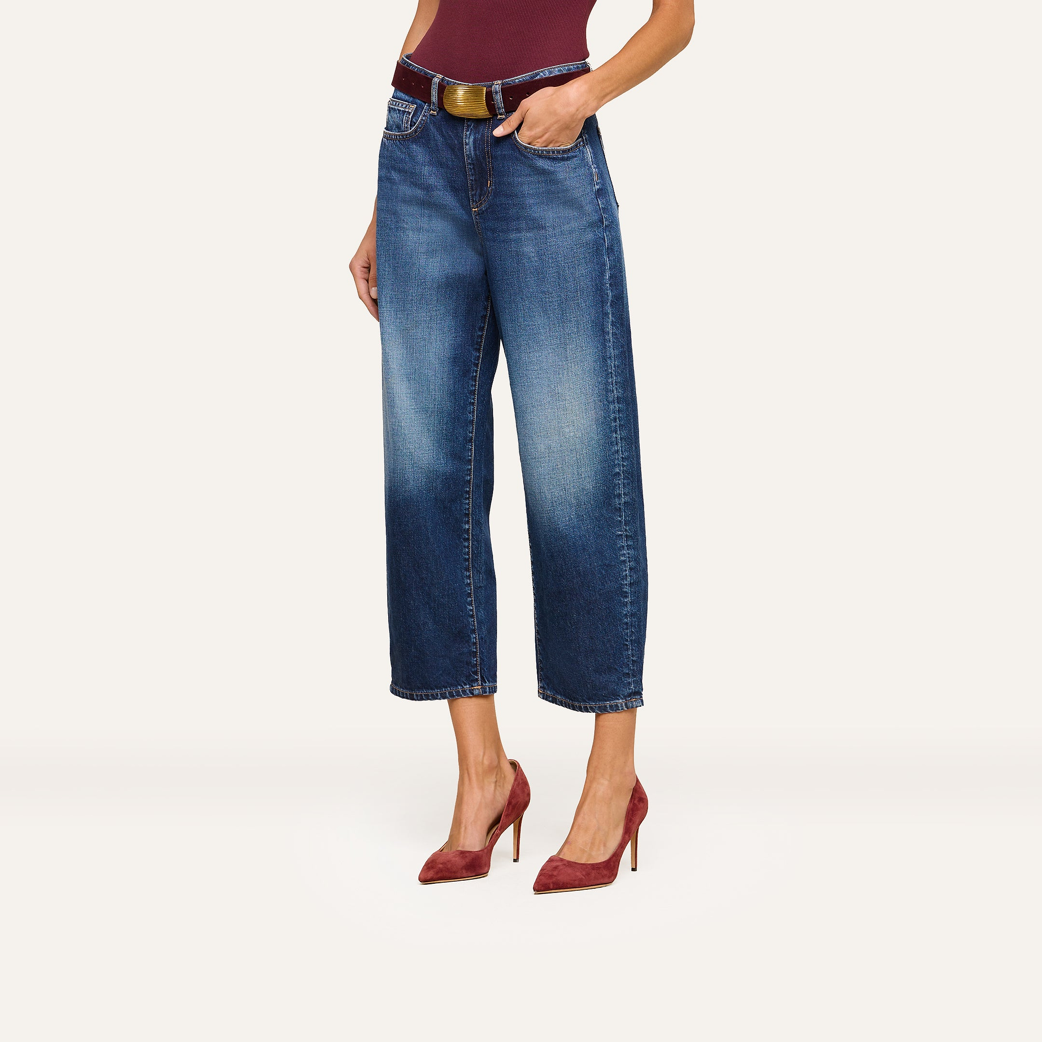 A woman wearing Mallis Bow-Leg Jeans in indigo wash, styled with a brown belt, a burgundy top, and matching burgundy high-heeled shoes, stands against a plain light background.