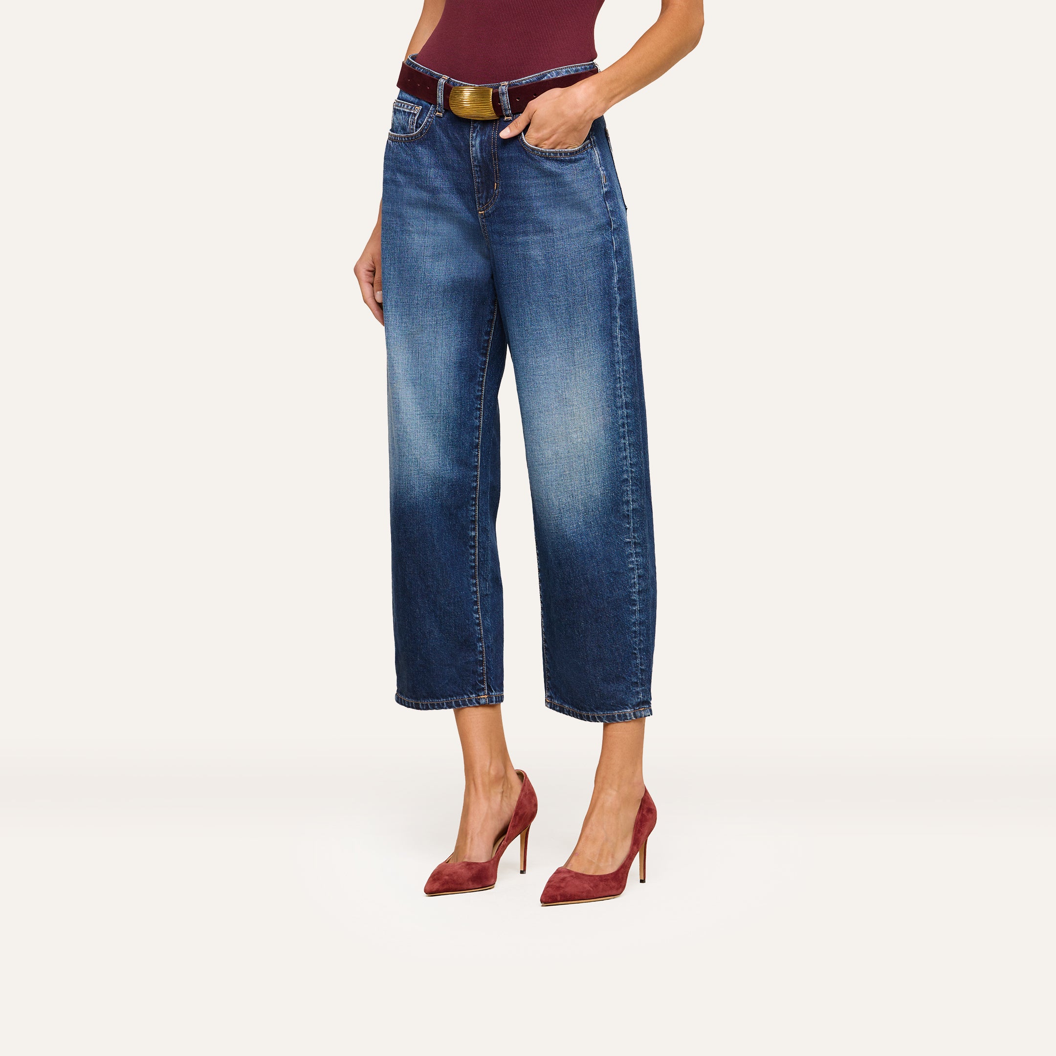 A woman wearing Mallis Bow-Leg Jeans in indigo wash, styled with a brown belt, a burgundy top, and matching burgundy high-heeled shoes, stands against a plain light background.
