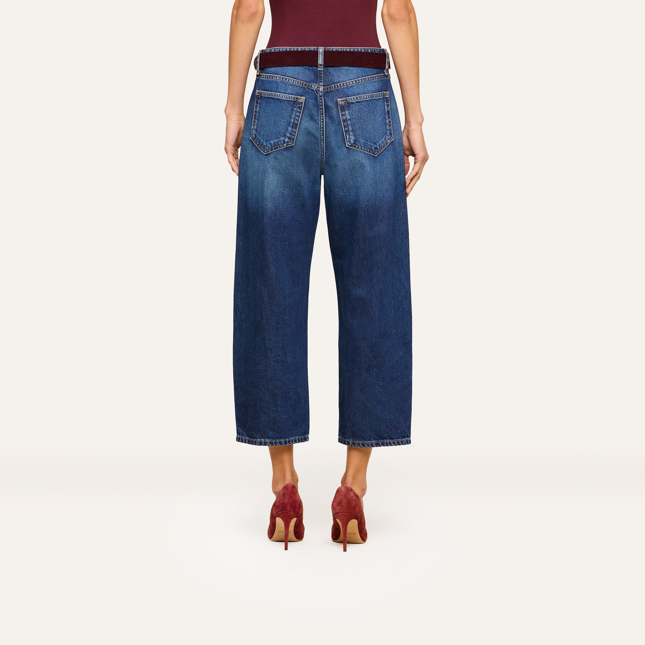 A person is shown from the back wearing the Mallis Bow-Leg Jean in an indigo wash paired with red high-heeled shoes. Their arms are relaxed at their sides, and the background is plain and light-colored.
