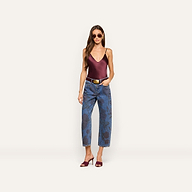 A woman wearing sunglasses, a burgundy satin camisole, Mallis Bow-Leg Jeans with a high-waisted pattern, a white belt, and burgundy heeled sandals stands against a plain light background.
