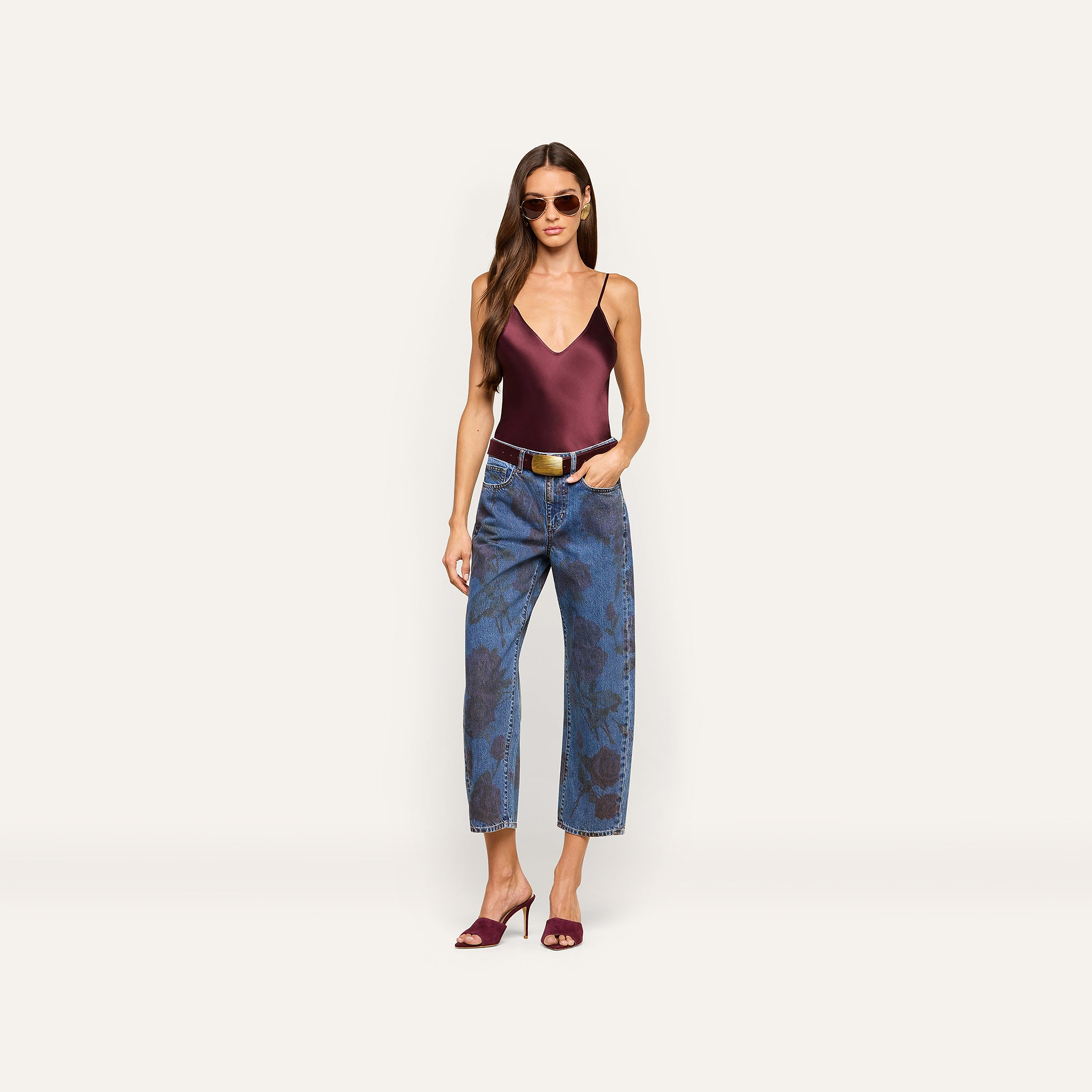 A woman wearing sunglasses, a burgundy satin camisole, Mallis Bow-Leg Jeans with a high-waisted pattern, a white belt, and burgundy heeled sandals stands against a plain light background.