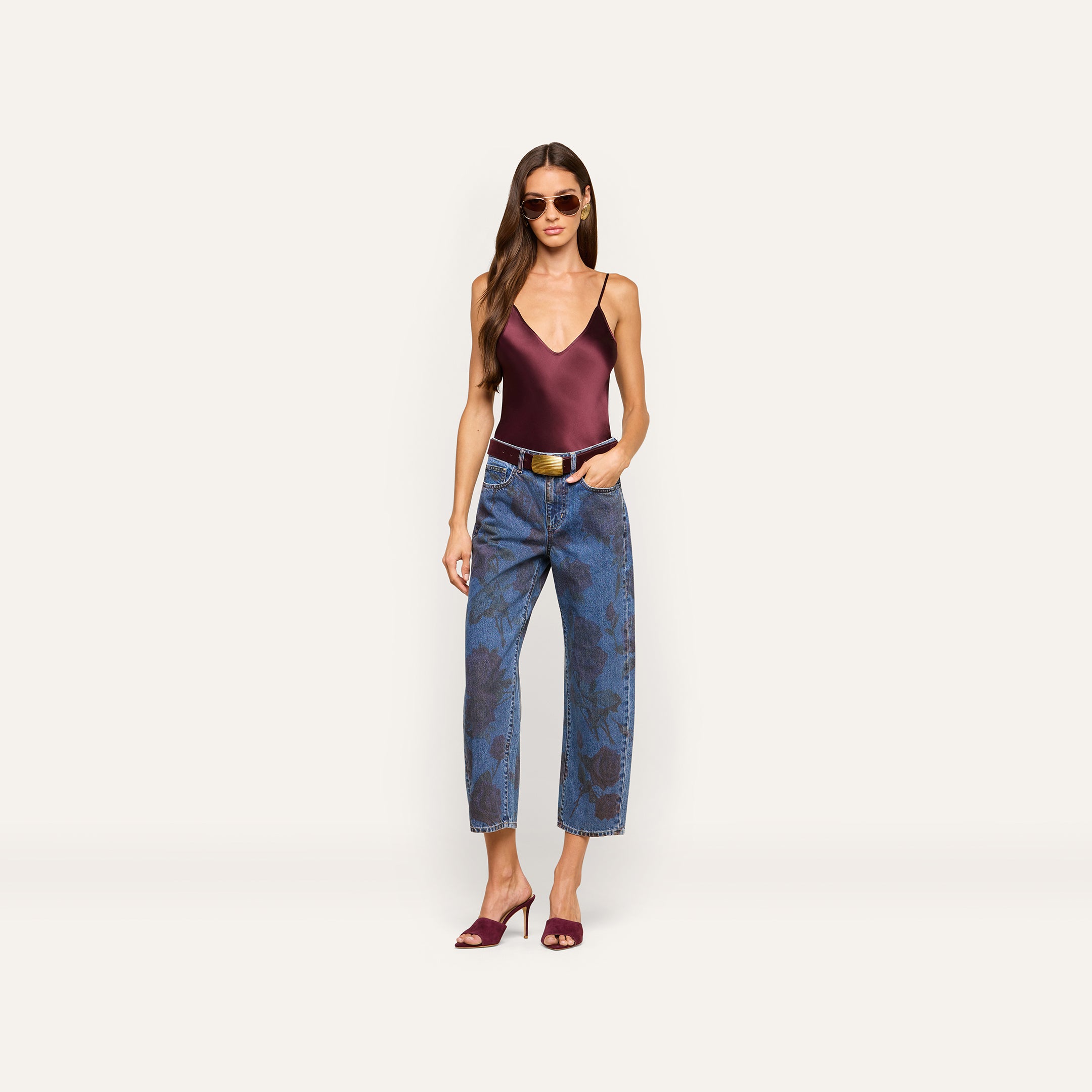 A woman wearing sunglasses, a burgundy satin camisole, Mallis Bow-Leg Jeans with a high-waisted pattern, a white belt, and burgundy heeled sandals stands against a plain light background.