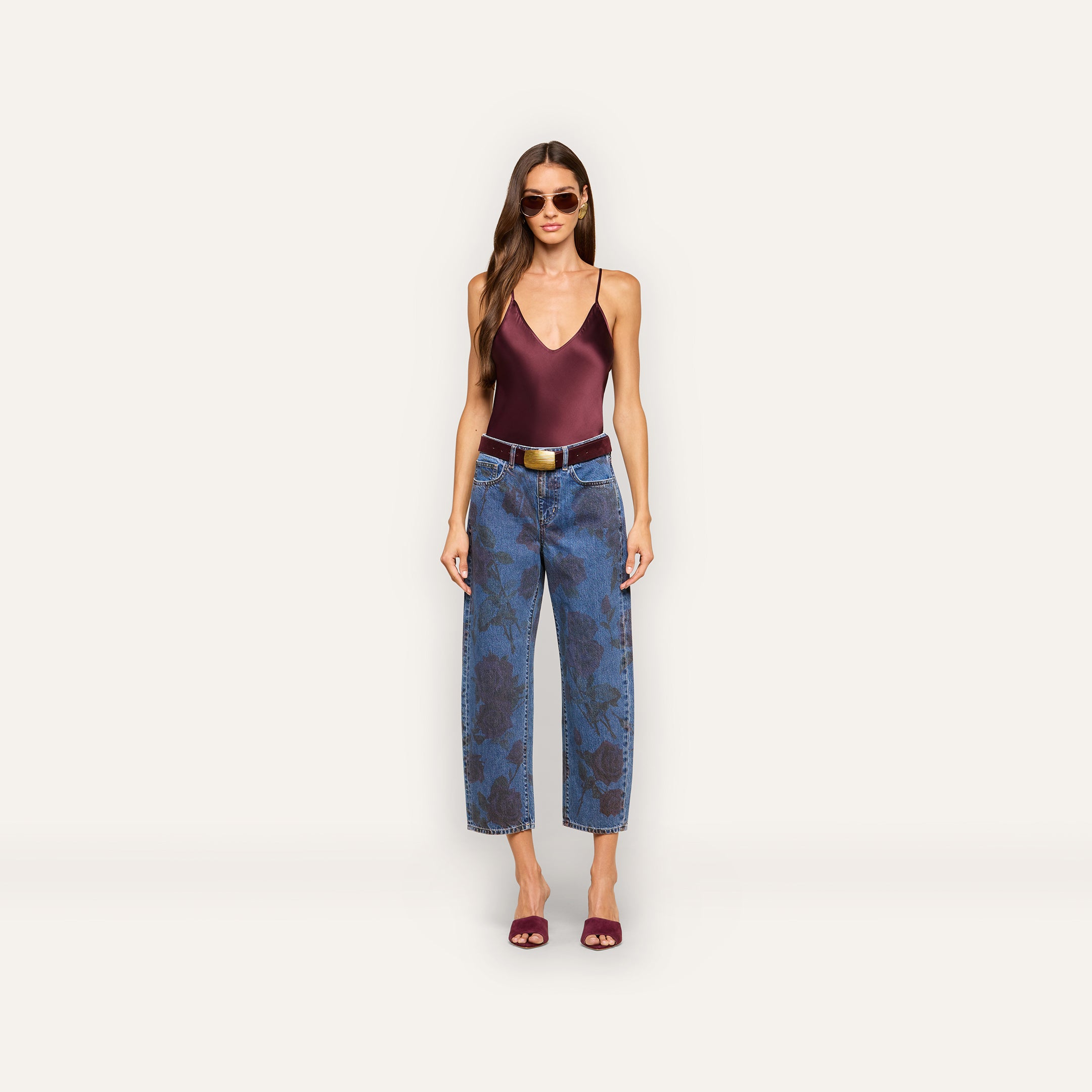 A woman wearing sunglasses, a maroon sleeveless top, the Mallis Bow-Leg Jean—a pair of blue high-waisted jeans with a dark rose print and wide-leg silhouette—a brown belt, and maroon heeled sandals stands against a plain light background.