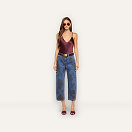 A woman wearing sunglasses, a maroon sleeveless top, the Mallis Bow-Leg Jean—a pair of blue high-waisted jeans with a dark rose print and wide-leg silhouette—a brown belt, and maroon heeled sandals stands against a plain light background.