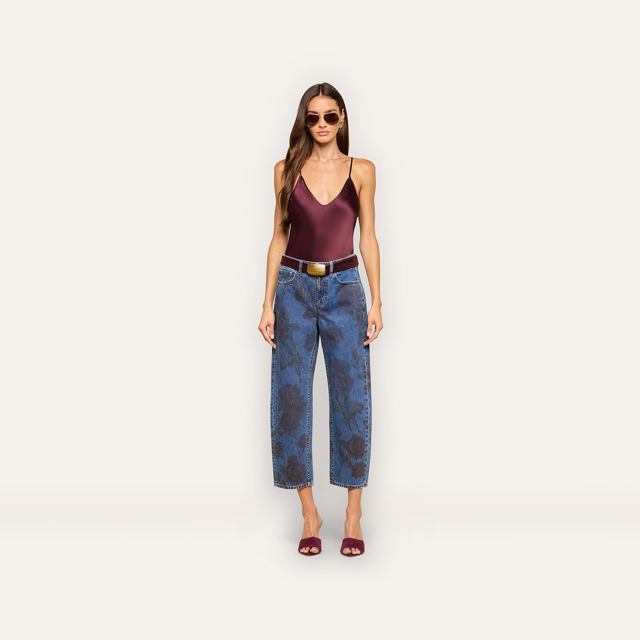 A woman wearing sunglasses, a maroon sleeveless top, the Mallis Bow-Leg Jean—a pair of blue high-waisted jeans with a dark rose print and wide-leg silhouette—a brown belt, and maroon heeled sandals stands against a plain light background.
